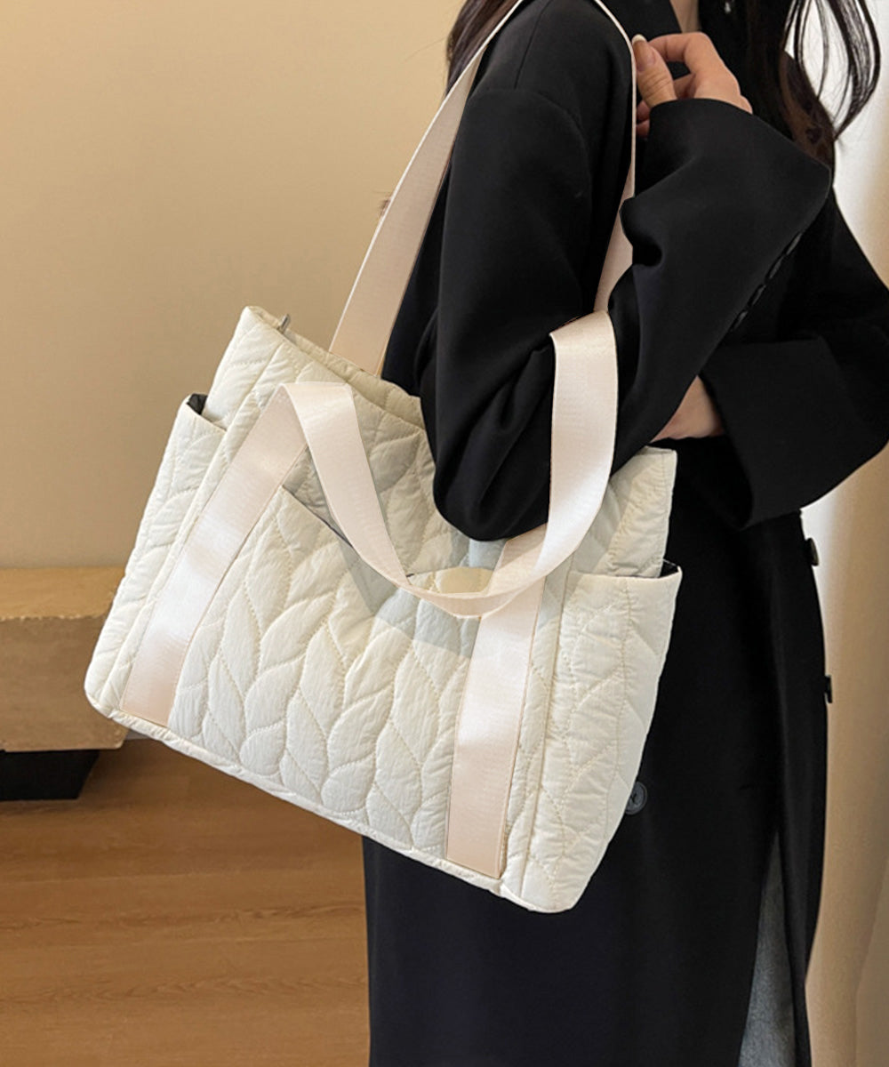 旅行や日常使いにぴったりの軽量キルティングトートバッグ。黒いコートを着た女性が白いバッグを肩にかけており、葉の模様が施されたデザインが特徴。幅広の持ち手で肩掛けしやすく、柔らかい素材で快適な使用感。シンプルかつエレガントなデザインで、どんなコーディネートにも合わせやすく、20~30代の日本人女性に最適なアイテム。